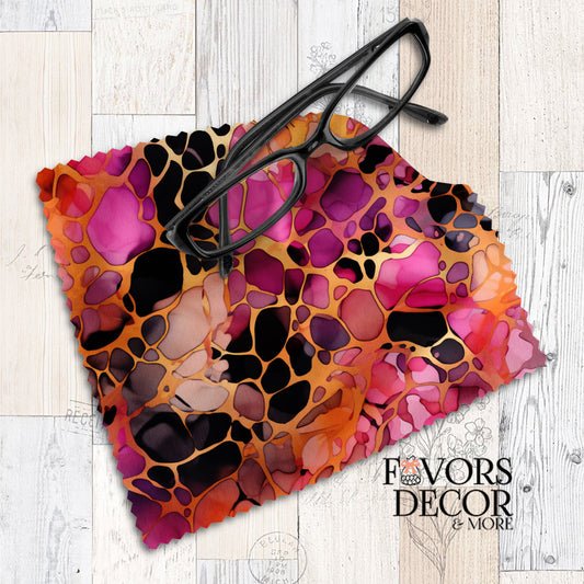Microfiber lens cleaning cloth with a vibrant berry-toned leopard print in pink, orange, and black, displayed with black eyeglasses on a wood surface.