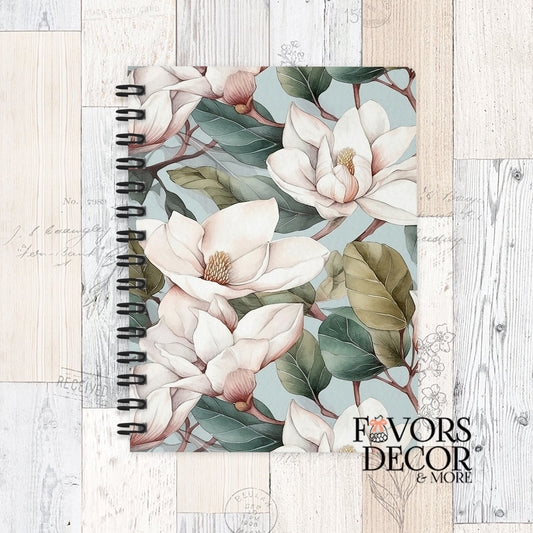 Spiral notebook with cream magnolia blossoms and green leaves on a soft blue background, displayed on a rustic wood surface with “Favors Decor &amp; More” logo.
