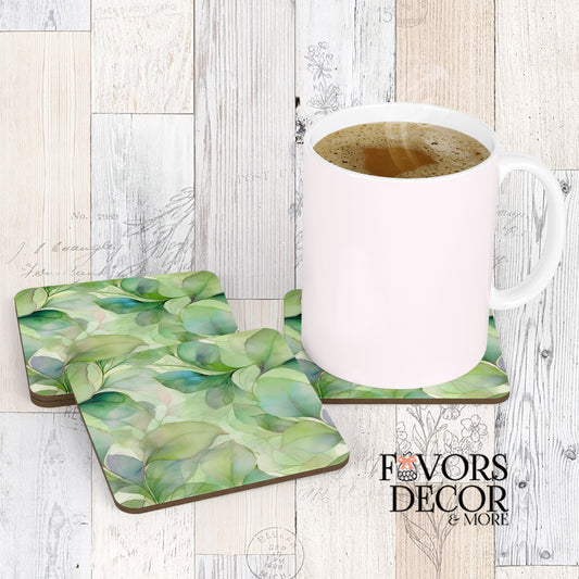 Two square coasters with a fluid green alcohol ink design featuring eucalyptus-like leaves in mint, sage, and subtle purple hues, with a white coffee mug sitting on one of them. The coasters are on a light wooden background.